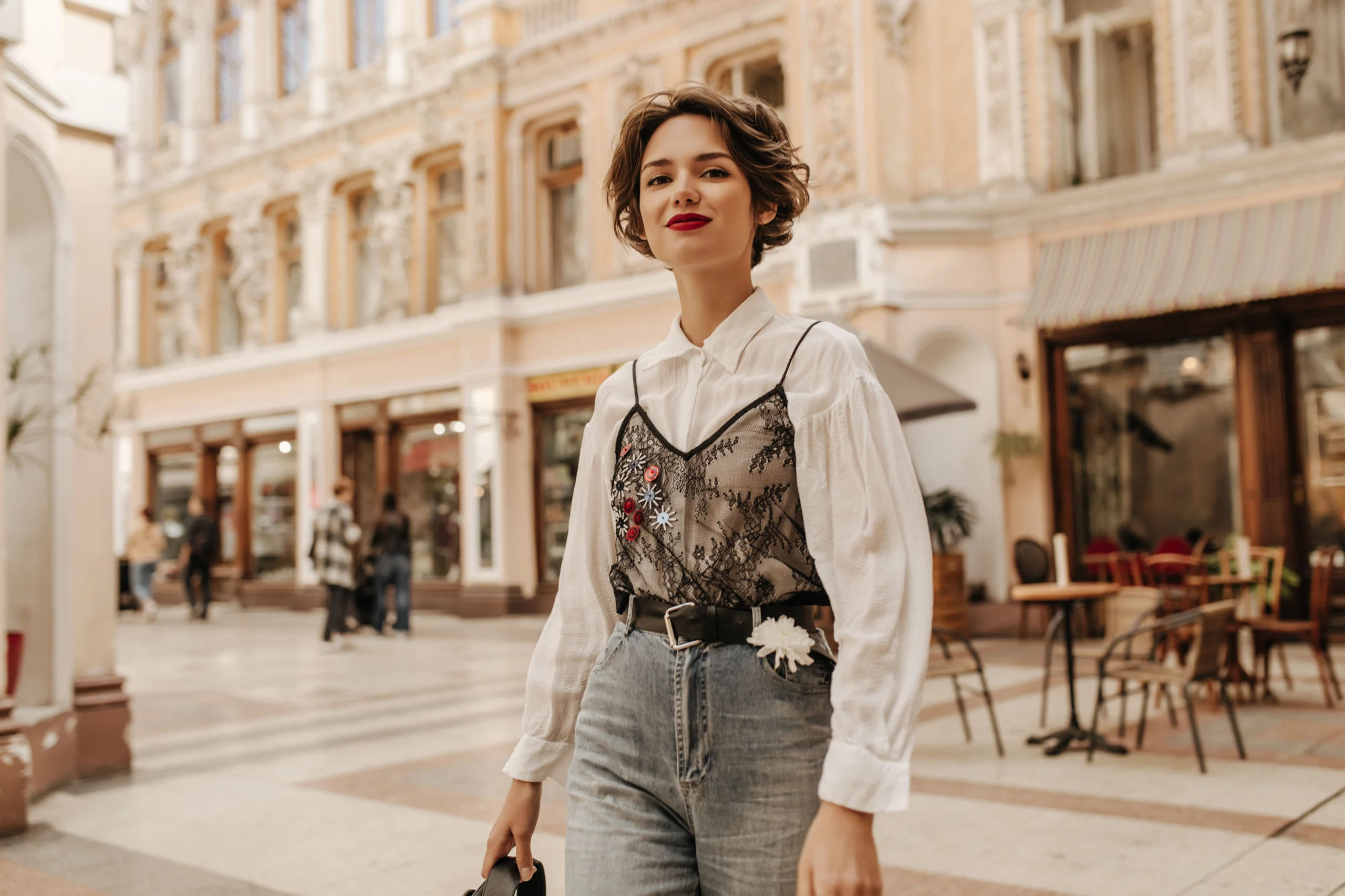hermosa mujer con cabello ondulado en jeans con cinturón y flor sonriendo en la calle señora fresca en blusa blanca con encaje posando en la ciudad